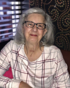 A woman is smiling at the camera. She's wearing glasses and a white shirt with red stripes. Her silver hair is pinned back. 