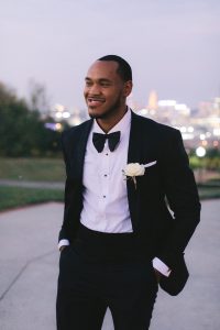 A man stands smiling in a tuxedo, his hands in his pockets.