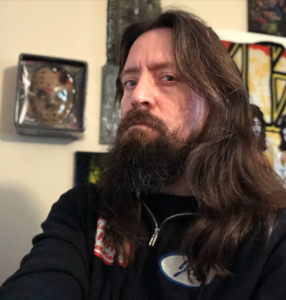 A man with long dark hair and a beard looks at the camera, unsmiling. He's wearing a black zip hoodie.
