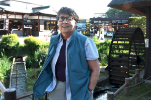 A woman with cropped grey hair and glasses, wearing a blue vest and short sleeve button down, stands in a garden